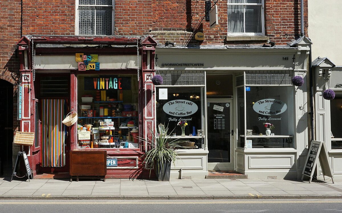 A colorful "Junk & Gems Vintage" shop sits beside "The Street Café and Coffee Bar" on a quiet brick-fronted street, with hanging baskets and a potted plant decorating the storefronts.