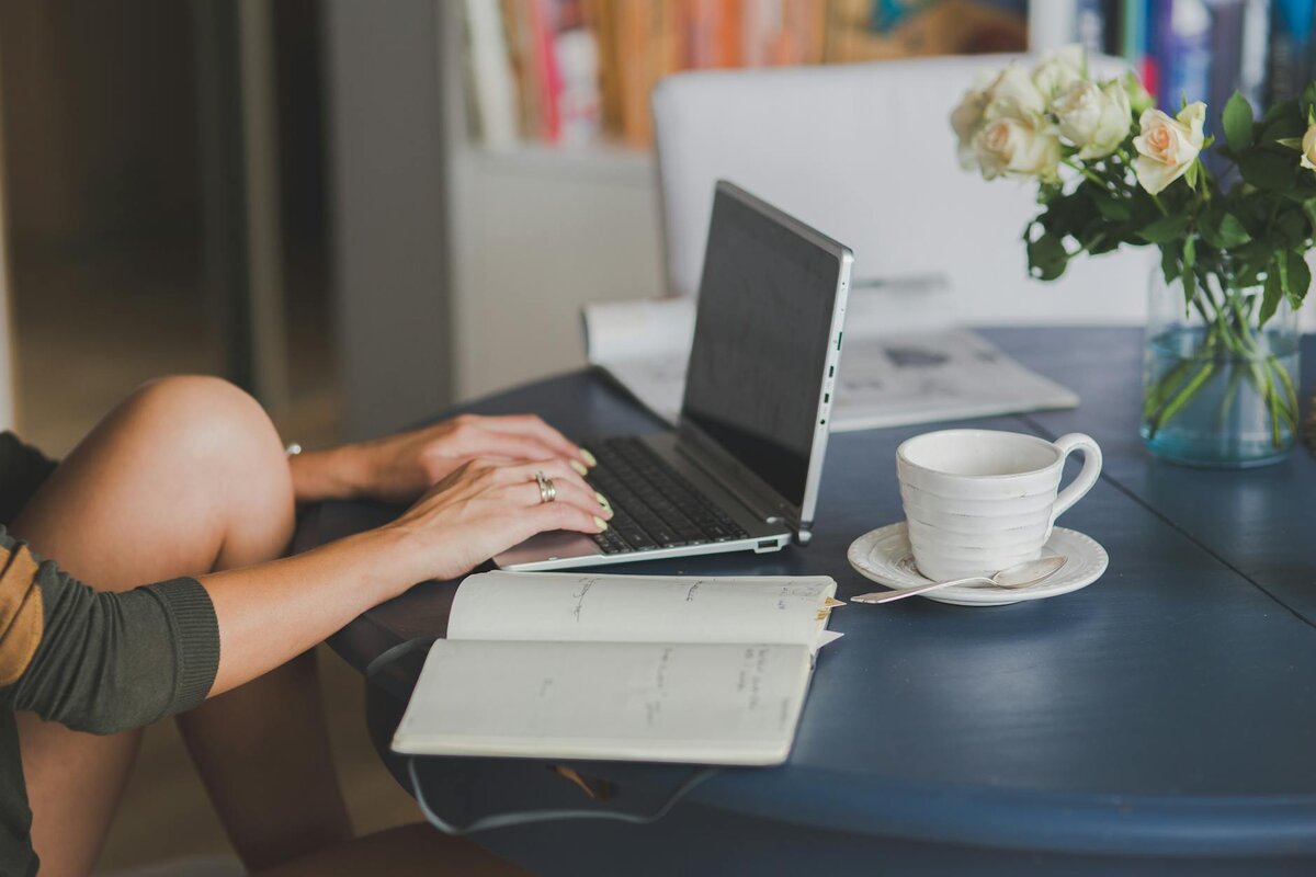 A close-up of a woman's hands typing on a laptop at a dark table, with an open notebook, a teacup, and a bouquet of roses nearby.