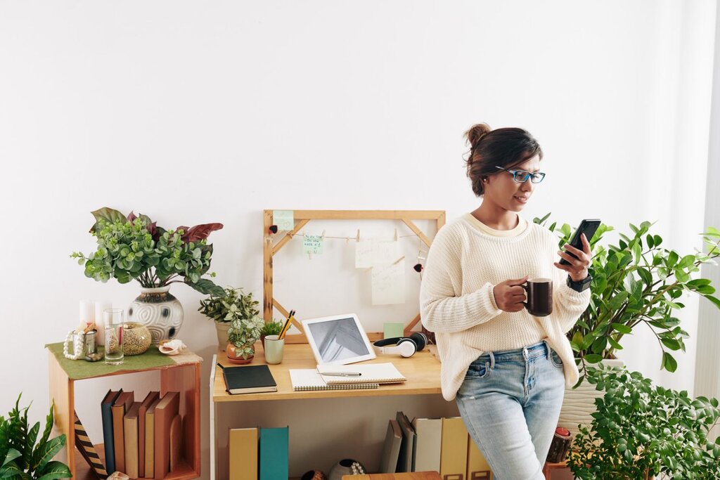 Woman wearing glasses stands holding a mug and looking at her phone, surrounded by plants and a desk setup.