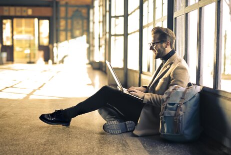 Man sits on the floor of a sunlit hallway using a laptop, with a bag beside him.