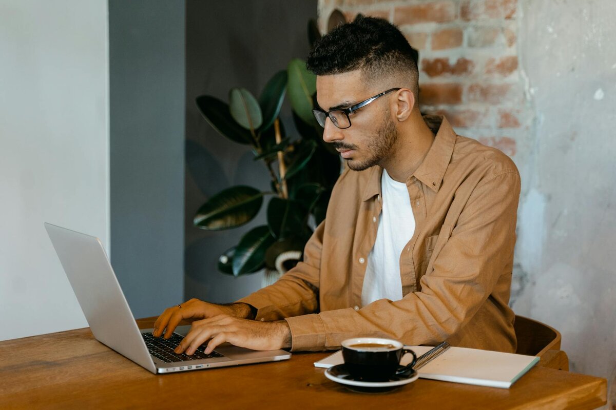 Man with glasses typing on a laptop at a wooden table, with a cup of coffee and a notebook beside him