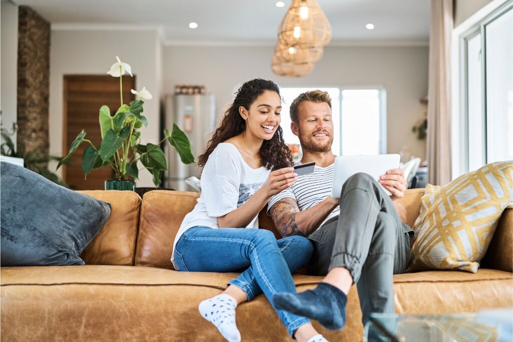 Smiling couple sit on a couch using a tablet and holding a credit card in a bright living room.