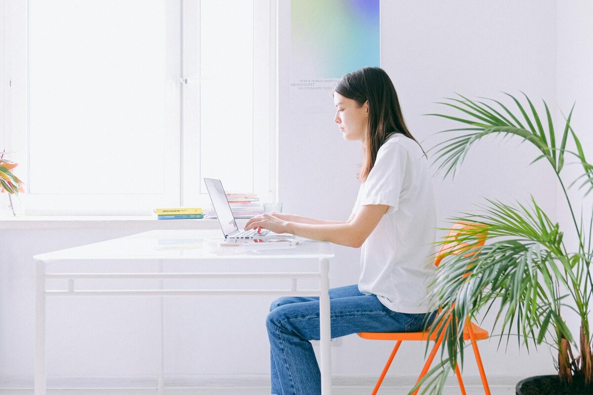 a woman types on a laptop at a white desk, sitting on an orange chair beside a leafy plant in a bright room