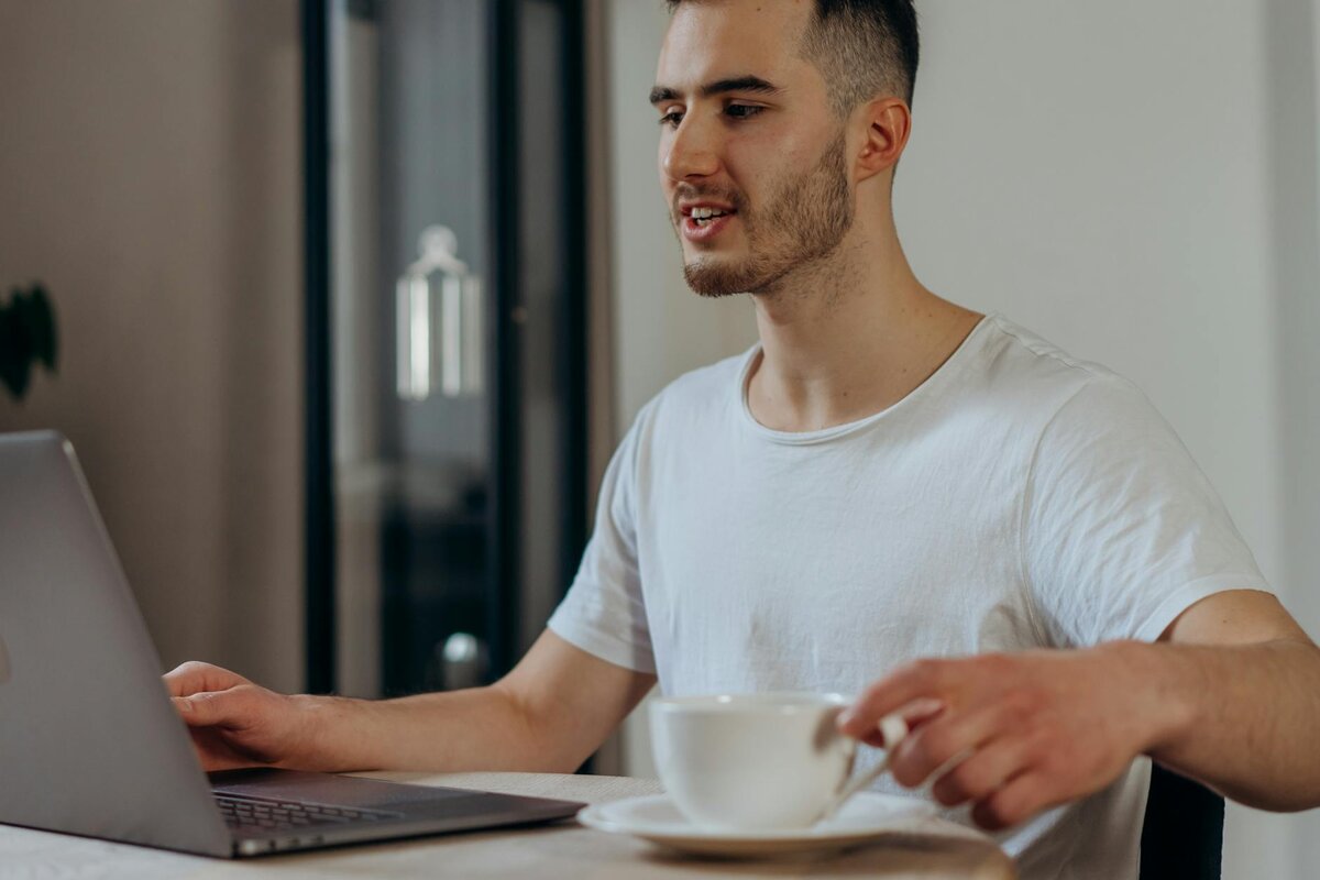 Man in white t-shirt using a laptop while holding a coffee cup at a table