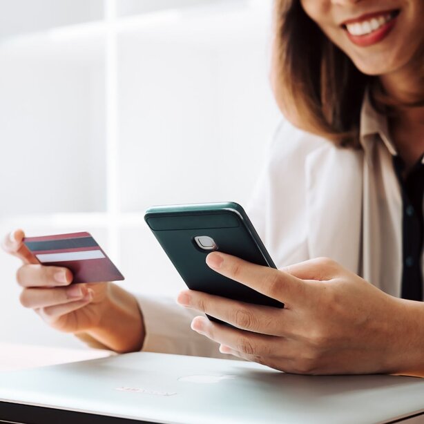 Smiling woman holding a credit card and using a smartphone, with a closed laptop on the table in front of her.