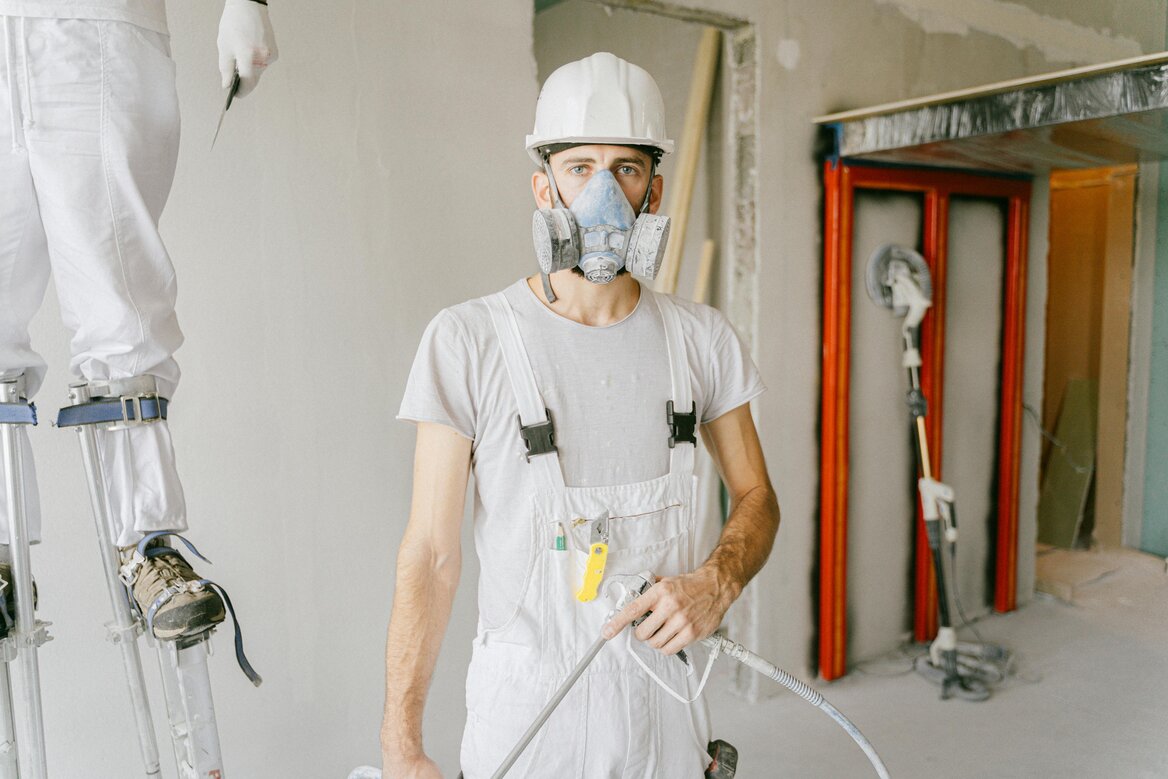 Worker in a hard hat, respirator, and overalls holding a spray tool, with a coworker on stilts beside him at a construction site.
