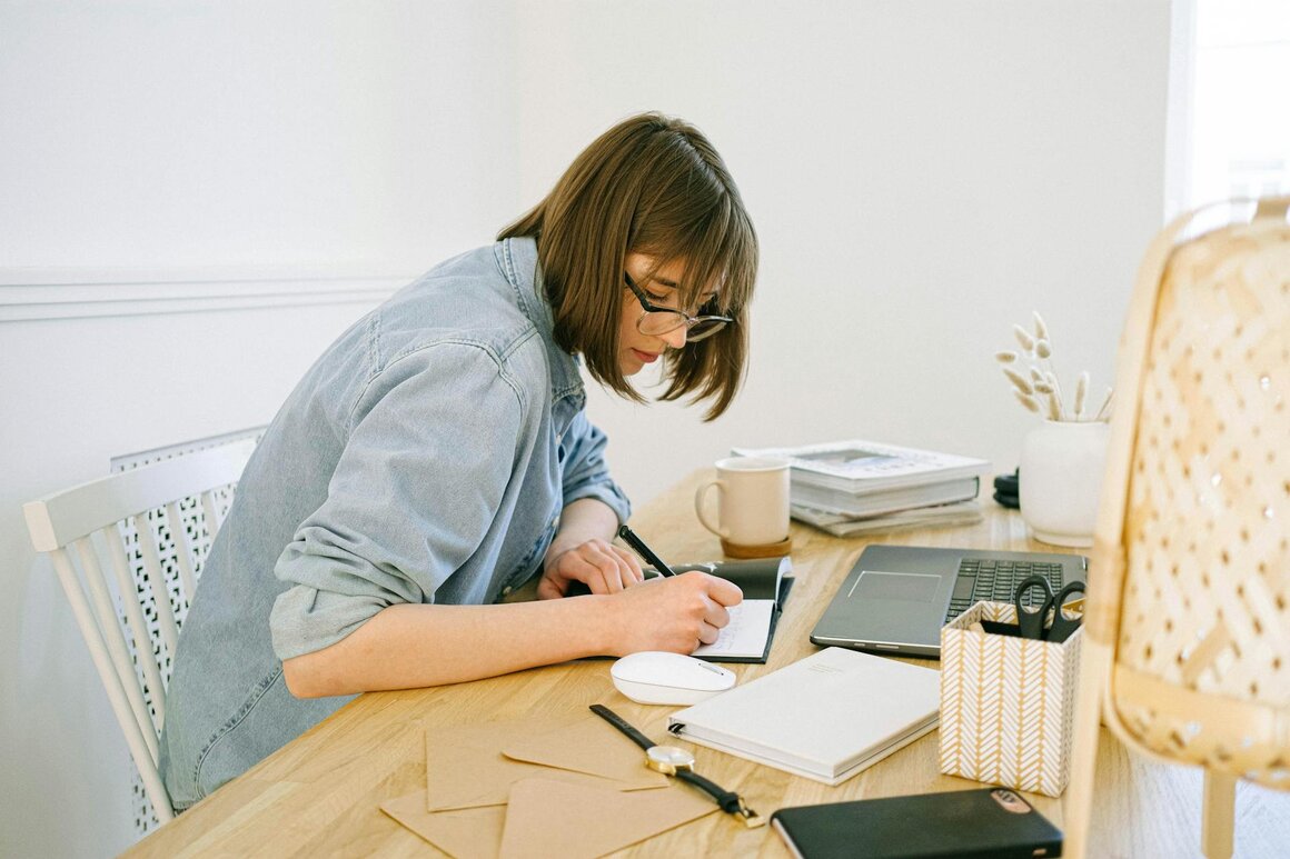 A woman on her desk, writing on her notebook in front of her laptop
