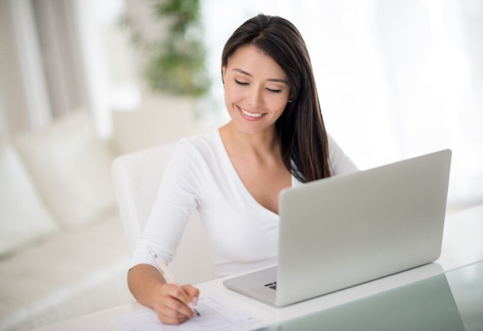 Woman writing notes at desk beside open laptop