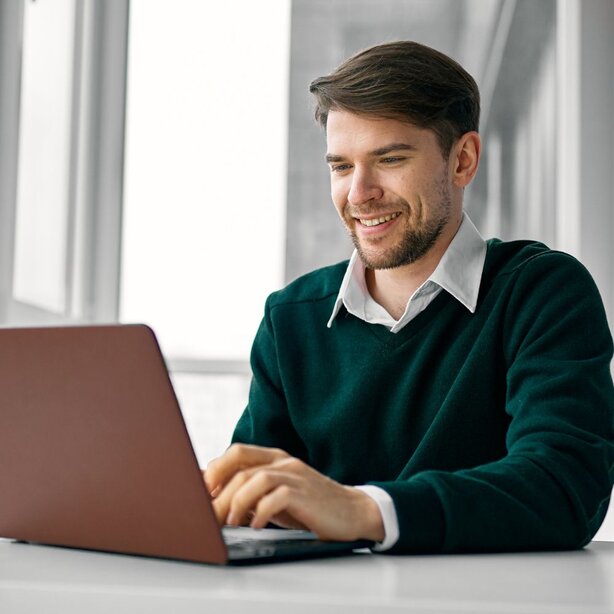 Smiling man typing on a laptop at a white desk near tall windows.