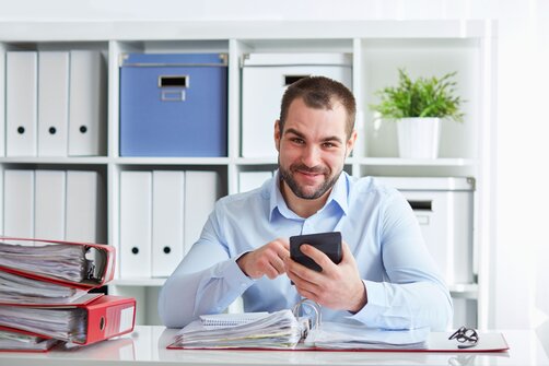 Smiling man sits at a desk holding a calculator, surrounded by binders and folders.