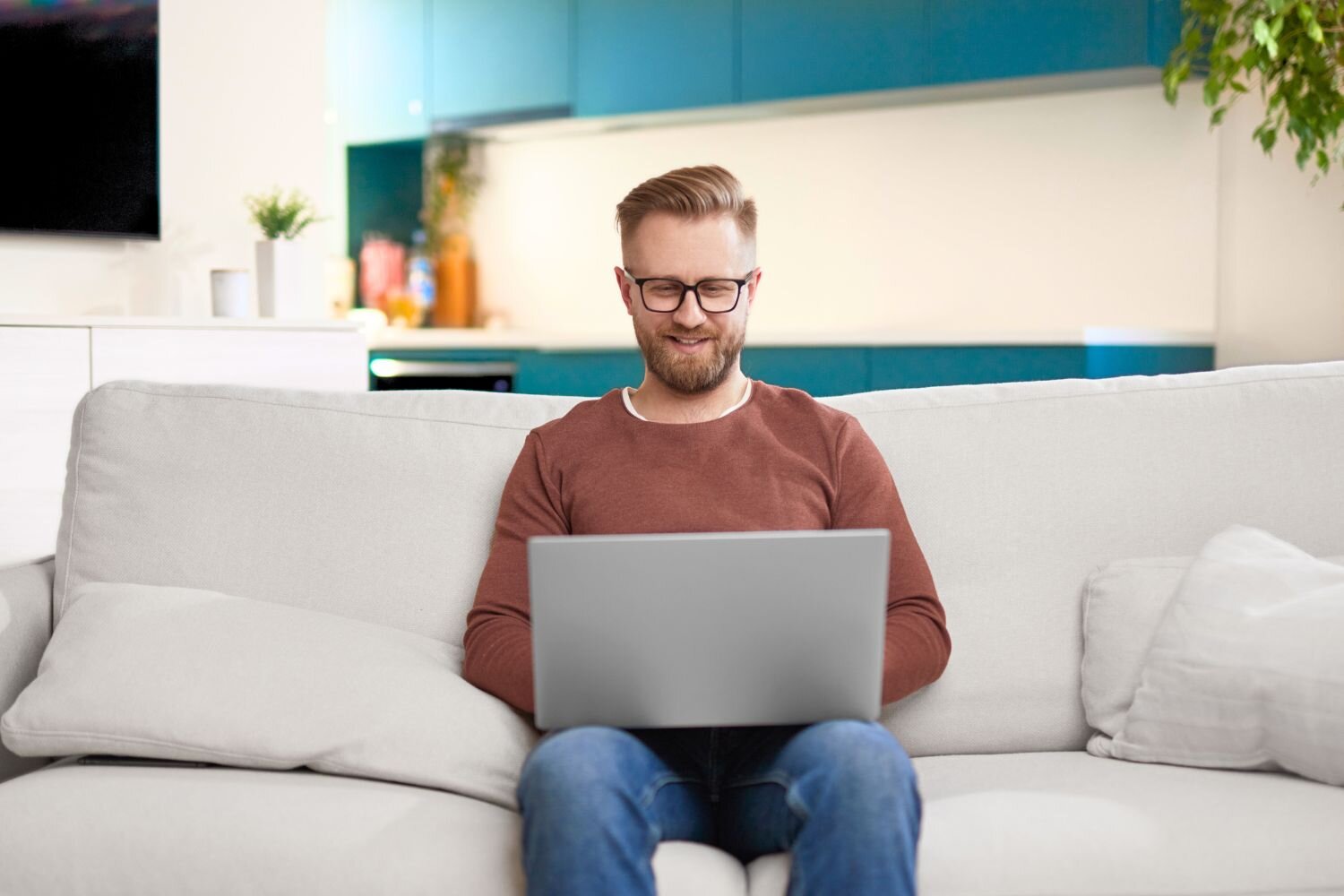 Smiling bearded man with glasses sitting on a couch using a laptop, with a kitchen visible behind him.