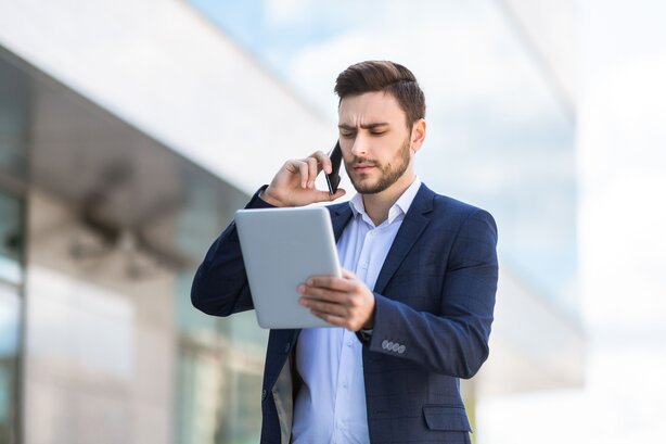 Man frowning while talking on the phone and looking at a tablet outdoors.