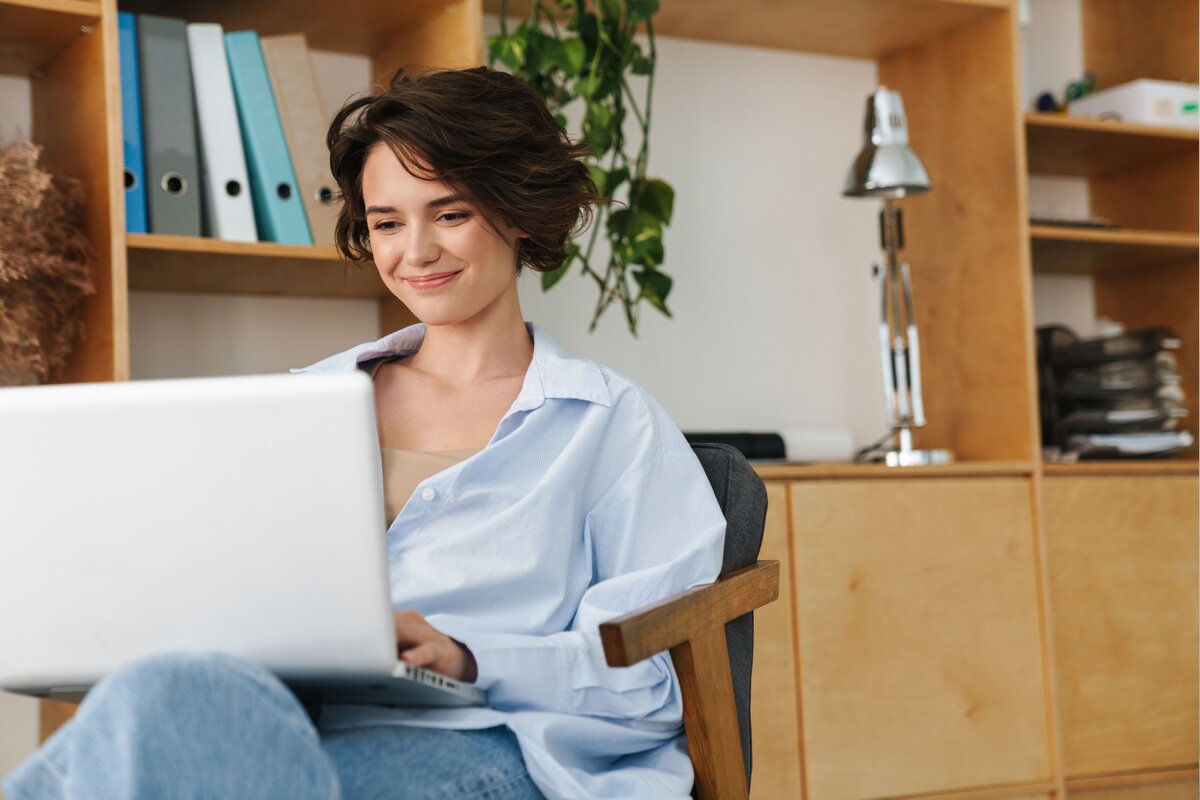 Smiling woman in an oversized shirt sitting in an armchair, typing on a laptop in front of wooden shelving with books and a desk lamp.
