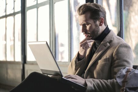 Man in a coat sits by a window with a thoughtful expression, looking at a laptop on his lap.