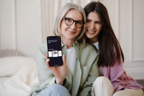 Two smiling women sitting close together, one holding up a phone displaying an app screen.