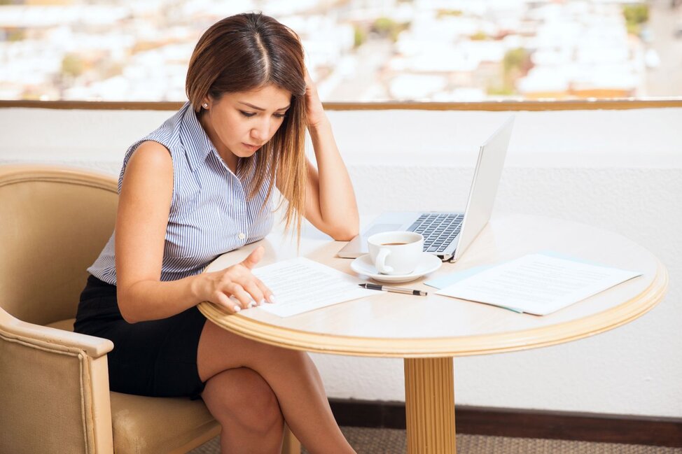 Woman sits at a small table reviewing paperwork with a laptop and coffee, looking concerned.