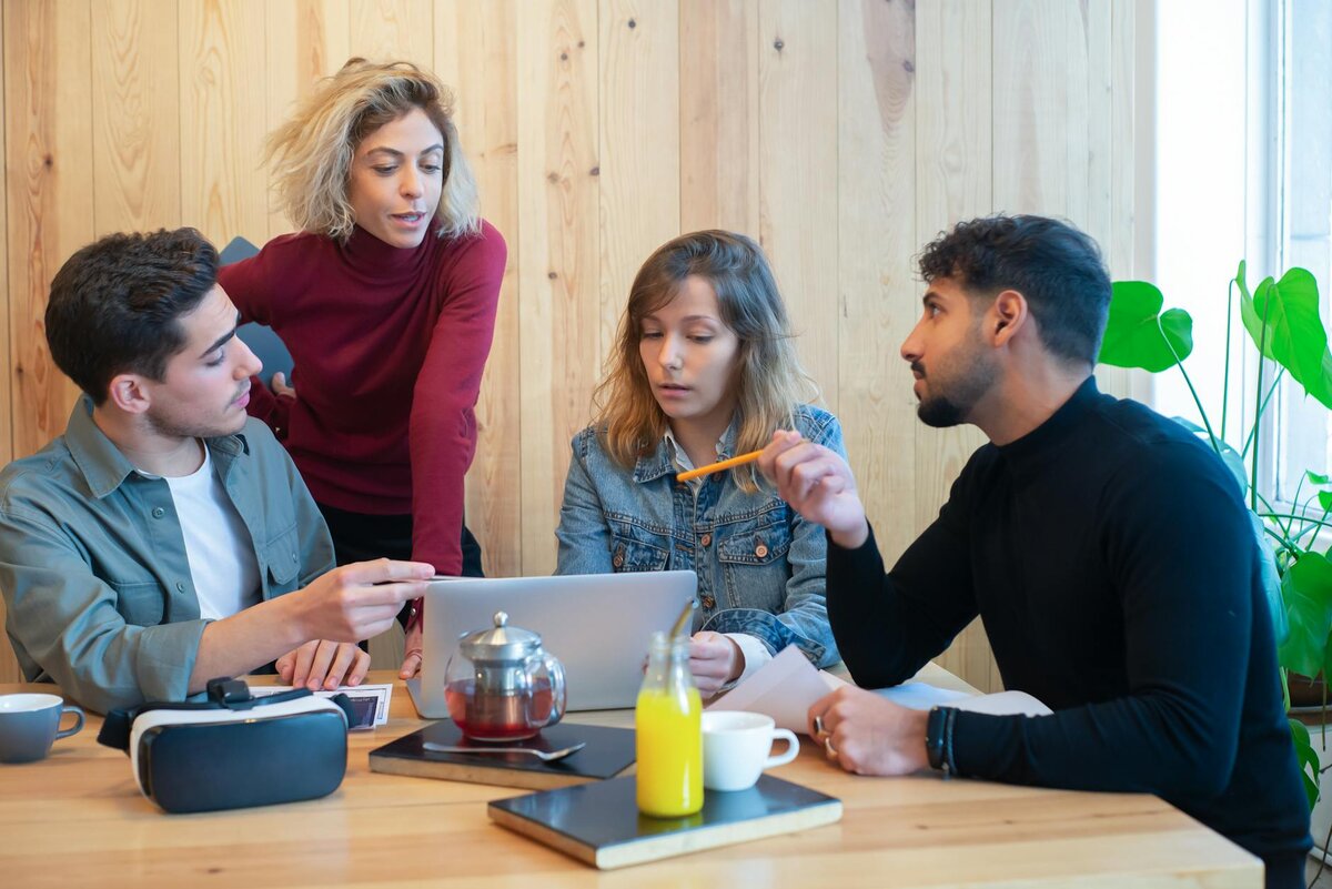 Four colleagues discuss work around a wooden table with a laptop, VR headset, and drinks, in a conversation.