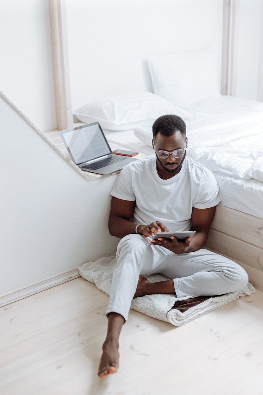Man in white clothing sits cross-legged on a wooden floor using a tablet, leaning against a bed in a bright minimalist room with a laptop and notebook behind him.