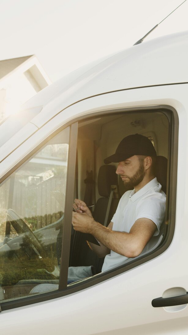 Man wearing a cap sits in the driver's seat of a white van writing on a notepad.