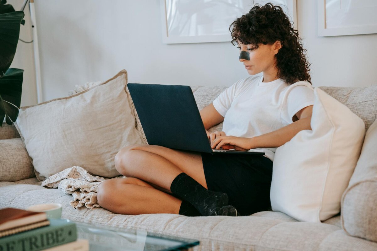 Woman relaxing on a sofa with a nose strip, using a laptop with cushions around her.
