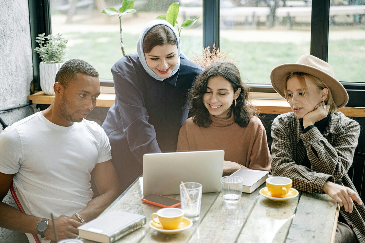 A diverse group of four friends gathers around a laptop at a café table with coffee cups and water glasses, smiling at the screen.