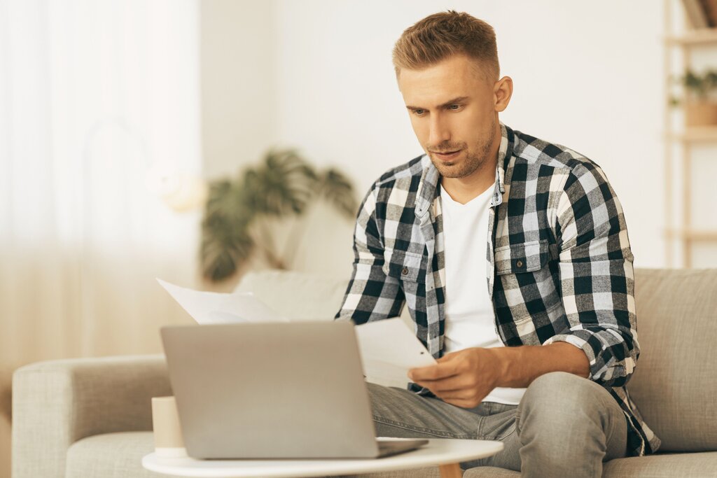 Man in flannel shirt reviewing paper while using home laptop