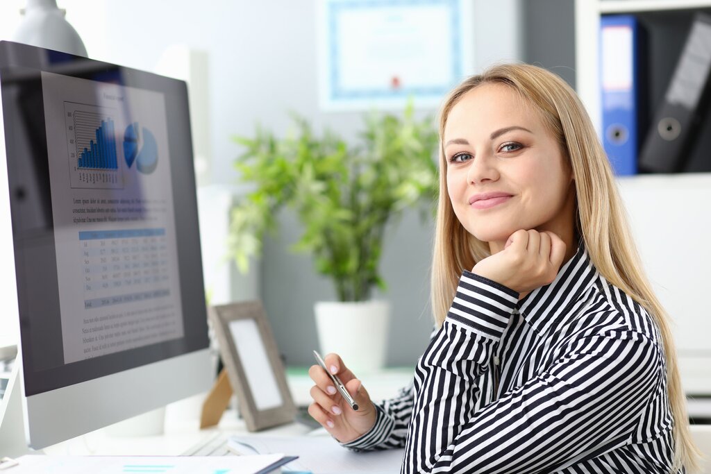 Woman smiling at the camera with her chin resting on her hand, a chart-filled monitor beside her.