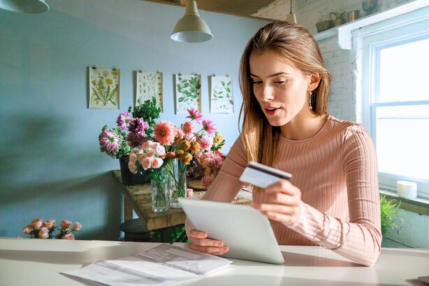 Woman holds a credit card while looking at a tablet at a kitchen counter with flowers nearby.