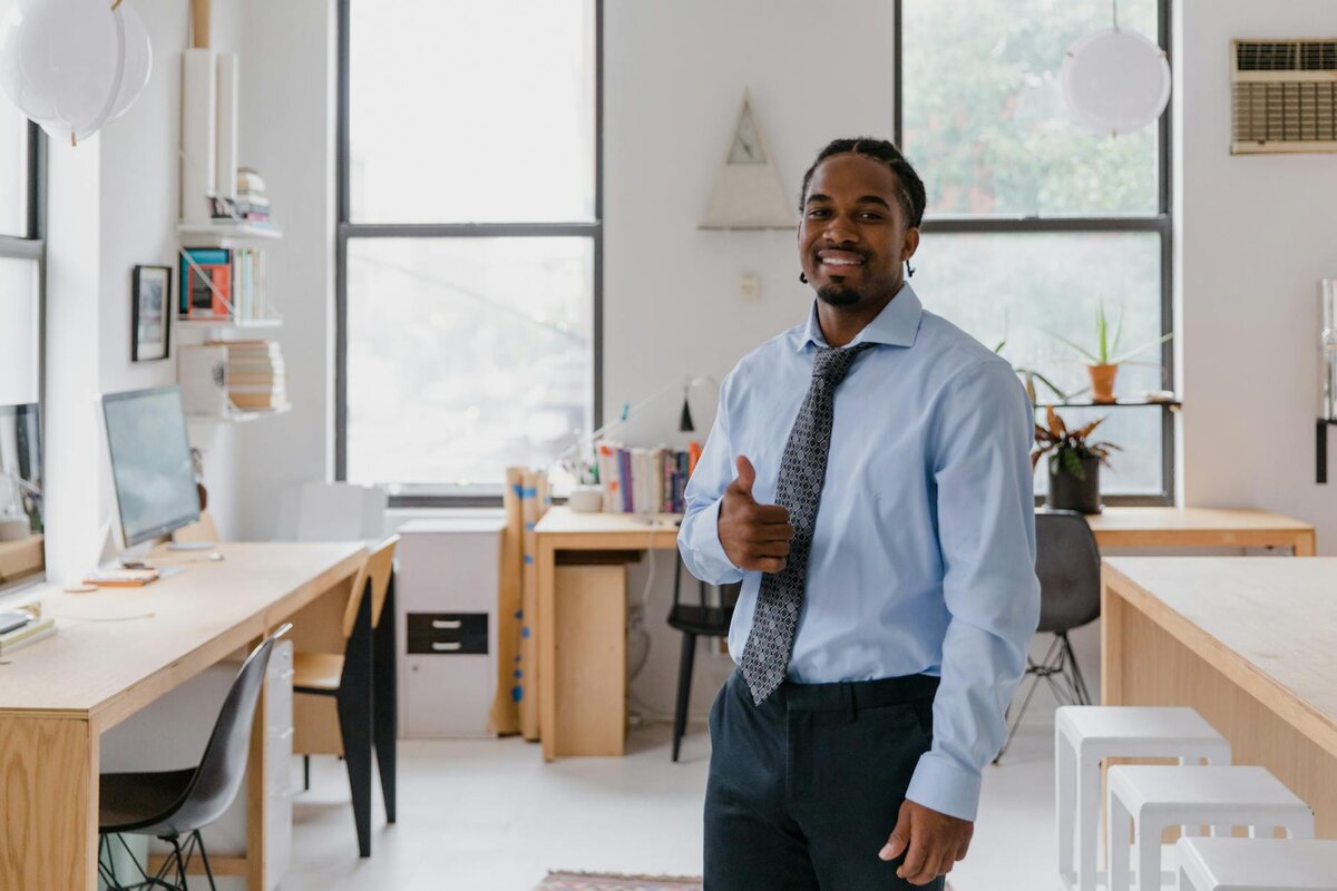 Smiling man in a dress shirt and tie giving a thumbs-up in a bright office space