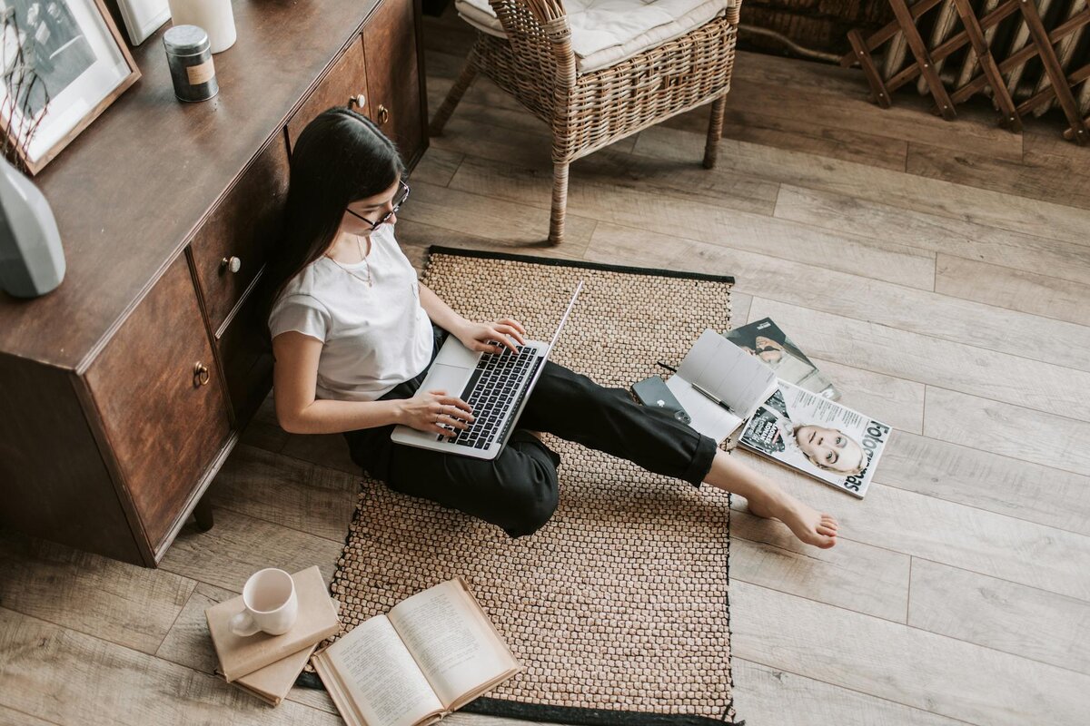 An overhead view of a woman sitting on the floor against a wooden cabinet, typing on a laptop surrounded by books, a notebook, and magazines on a woven rug.