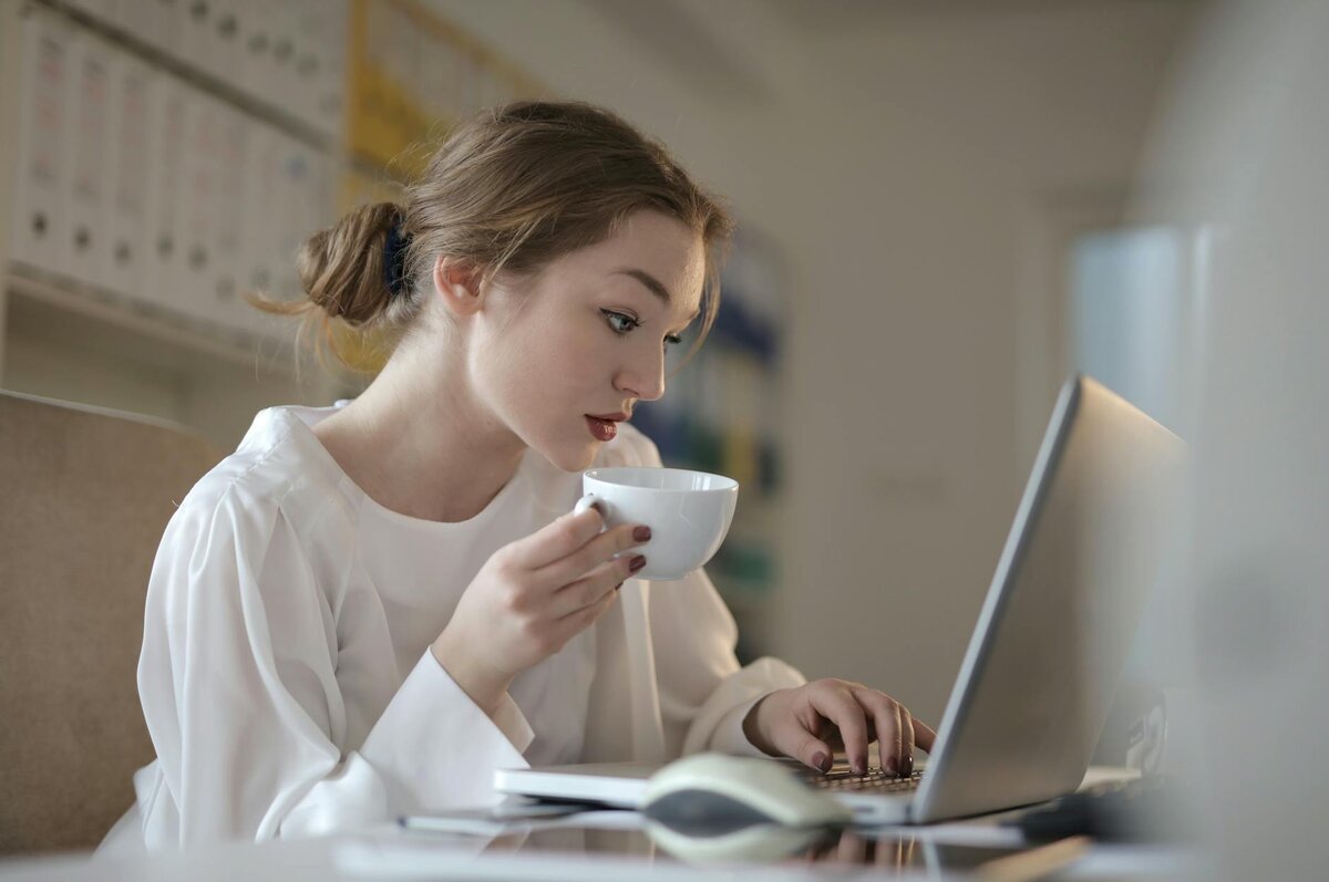woman in a white blouse sips from a cup while typing on a laptop at her desk, with binders visible in the background.