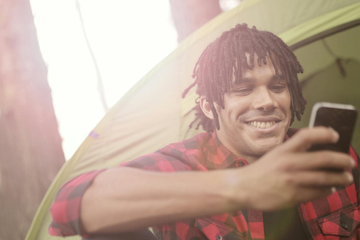 Smiling man with dreadlocks using smartphone inside green camping tent
