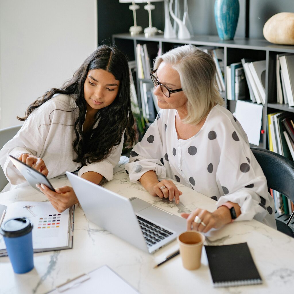 Two women sit together at a table reviewing a laptop and tablet, with notebooks, coffee cups, and a bookshelf in the background.