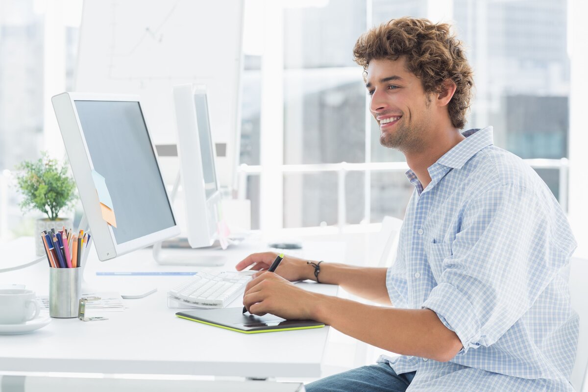 Smiling man working at a desk with two monitors and a graphics tablet in a bright office.